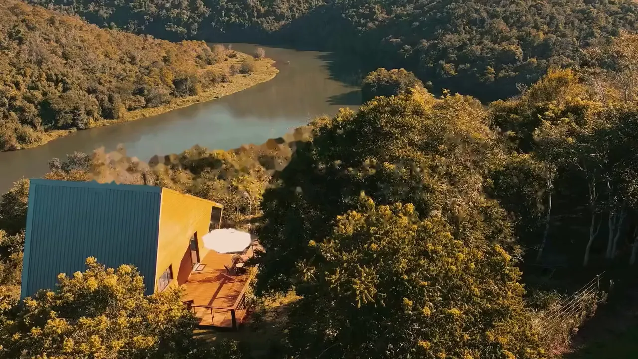 Cabana de madeira e vidro no alto da encosta, com o vale e a mata ao redor, vista de drone